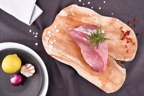 Surowa szynka wieprzowa EKO 500g na drewnianej desce z rozmarynem, solą, chili, cytryną, czosnkiem i cebulą